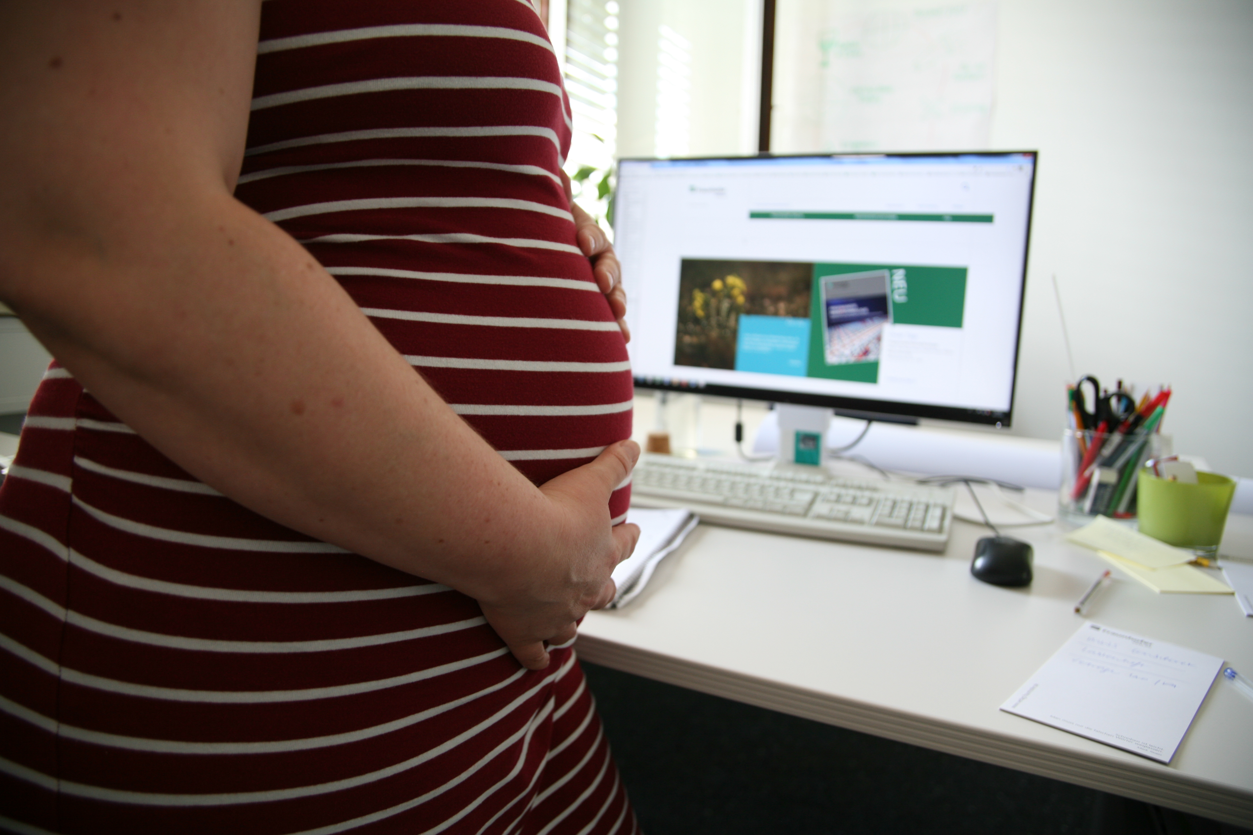 Der Babybauch einer werdenden Mama die vor ihrem Schreibtischsteht. Sie faltet die Hände über dem Bauch.