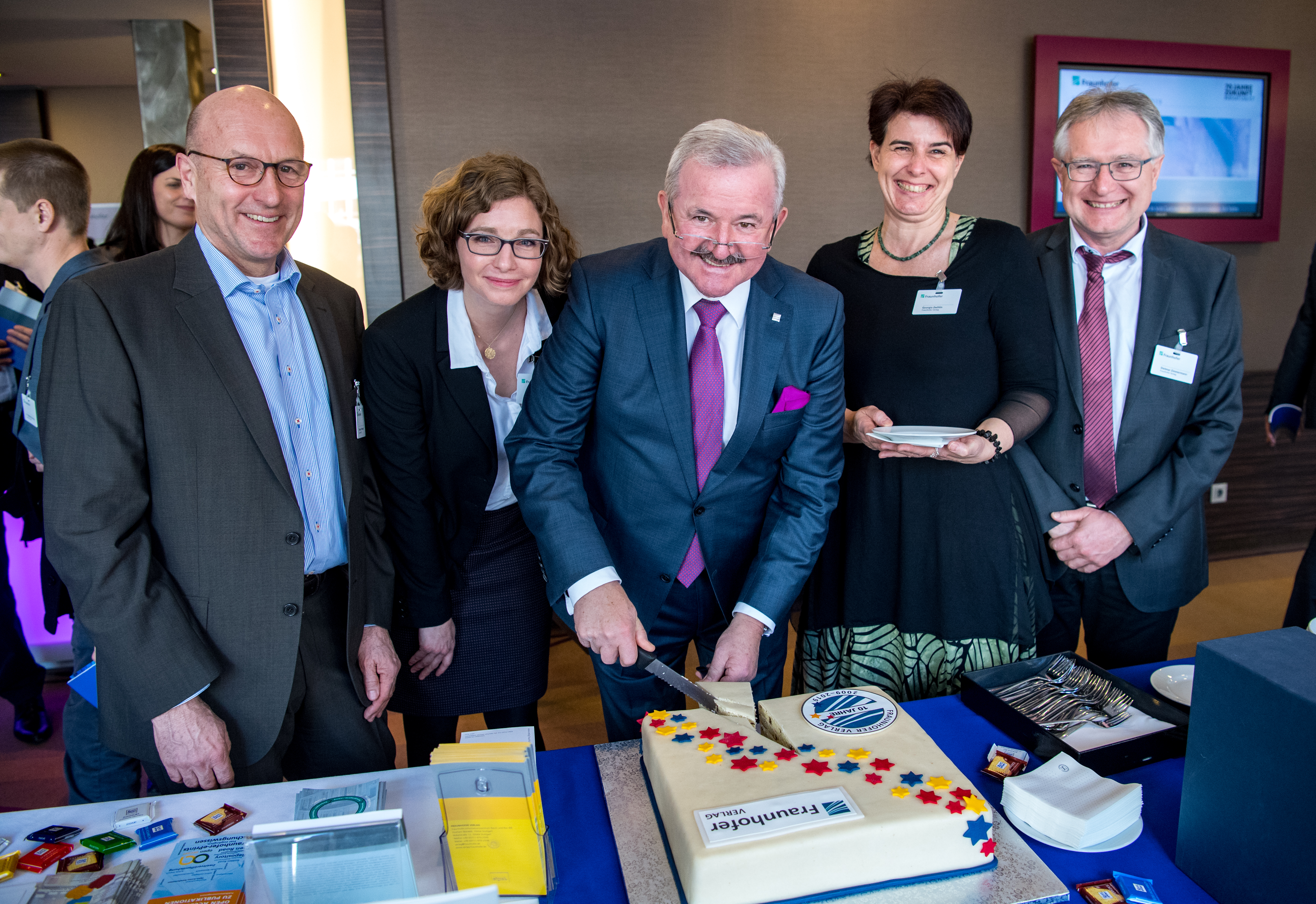 Zum 10-jährigen Jubiläum des Fraunhofer Verlag schneidet Reimund Neugebauer eine Torte an und blickt dabei freundlich in die Kamera.