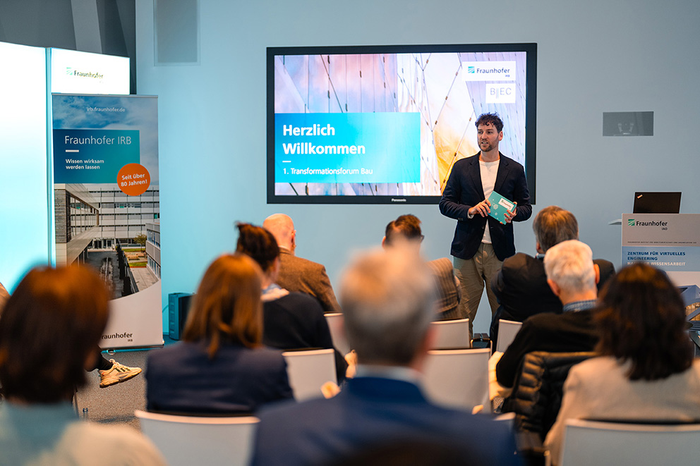 Ein Mann steht vor einem Publikum und heißt alle zum 1. Transformationsforum Bau 2024 willkommen.